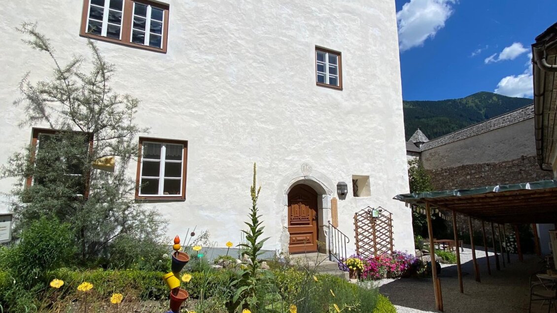 Ein charmantes weißes Gebäude mit einem einladenden Eingang. Im Vordergrund blühen gelbe Blumen und es gibt eine bunte Figur im Garten. | © Naturpark Sölktäler Julia Aigner
