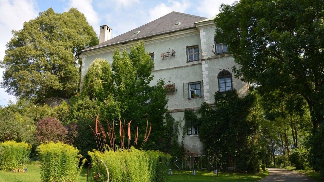 Ein historisches Gebäude, umgeben von üppigem Grün und bunten Pflanzen. Der Himmel ist teilweise bewölkt und verleiht der Szene eine ruhige Atmosphäre. | © TVB Murau