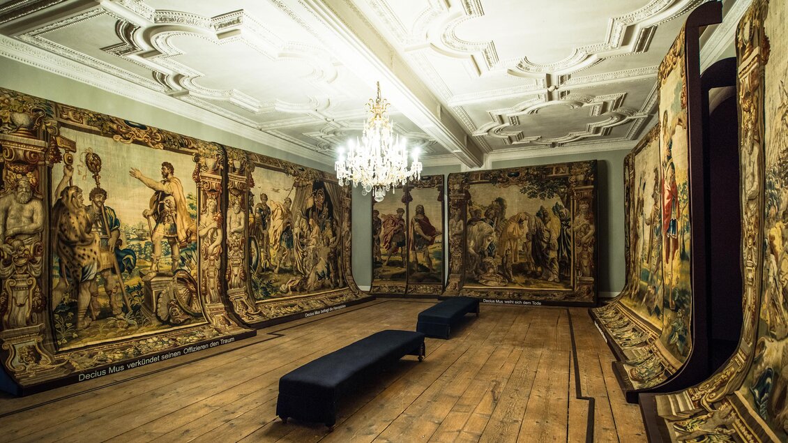 Ein eleganter Raum mit kunstvollen Wandteppichen und einem prachtvollen Kronleuchter. In der Mitte steht eine Bank auf einem Holzfußboden. | © TVB Murau