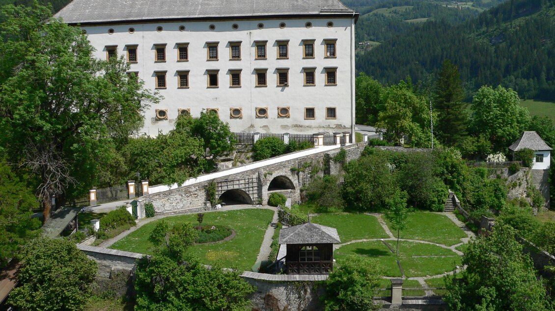 Ein großes, historisches Gebäude umgeben von grünen Bäumen und Gärten. Im Hintergrund sind sanfte Hügel und ein blauer Himmel zu sehen. | © TVB Murau