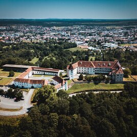 An impressive view of a historic castle, surrounded by green forests and a city in the background. The architecture is distinctive and well-preserved. | © Patrick Schadler | shutterstock.com