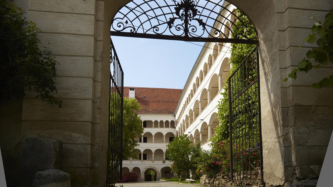 Ein schöner Innenhof mit historischen Gebäuden und grünen Pflanzen. Durch ein schmiedeeisernes Tor sieht man einen klaren Himmel. | © Manfred Donauer