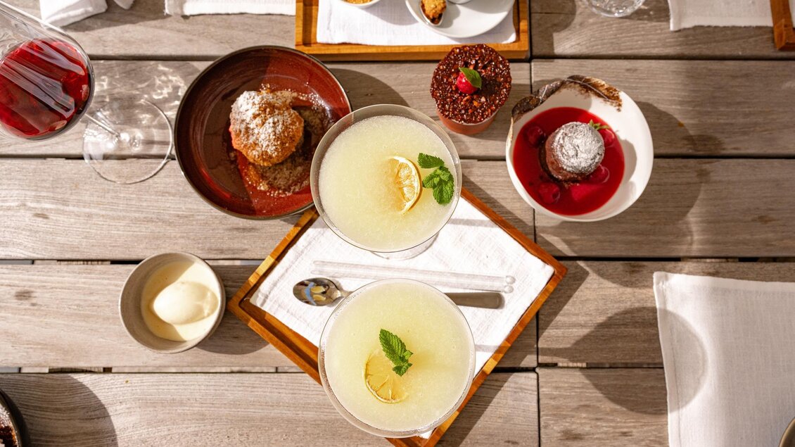 Zwei Cocktails mit Limette und Minze auf einem Tablett, umgeben von verschiedenen Desserts. Der Tisch ist aus Holz und gut dekoriert. | © Rene Strasser Fotografie