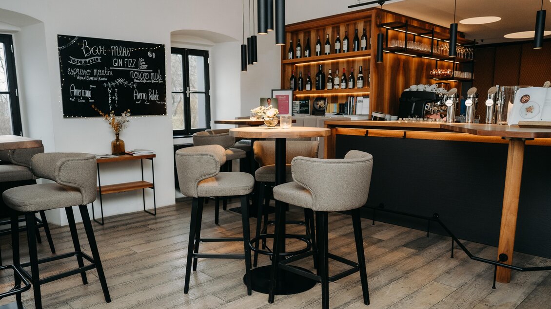 Eine moderne Bar mit eleganten Stühlen und einer gut sortierten Theke. An der Wand hängt eine Tafel mit Angeboten. | © Hutter