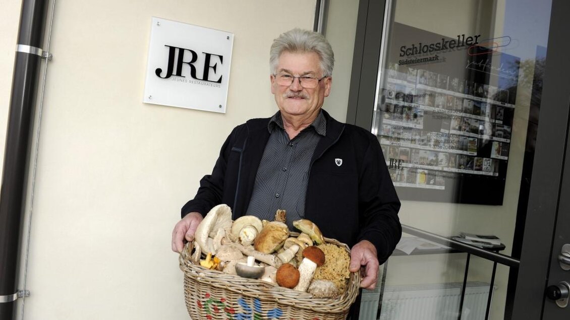 Ein älterer Mann steht vor einem Eingang und hält einen Korb mit verschiedenen Pilzen. Er trägt eine dunkle Jacke und lächelt freundlich in die Kamera. | © Schlosskeller Südsteiermark