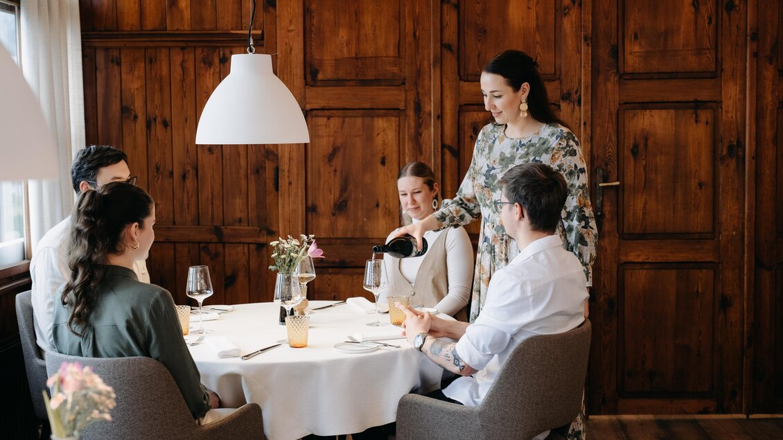Eine elegante Restaurantatmosphäre mit einem Tisch, der für vier Personen gedeckt ist. Eine Kellnerin serviert Getränke an die Gäste. | © Schlosskeller Südsteiermark