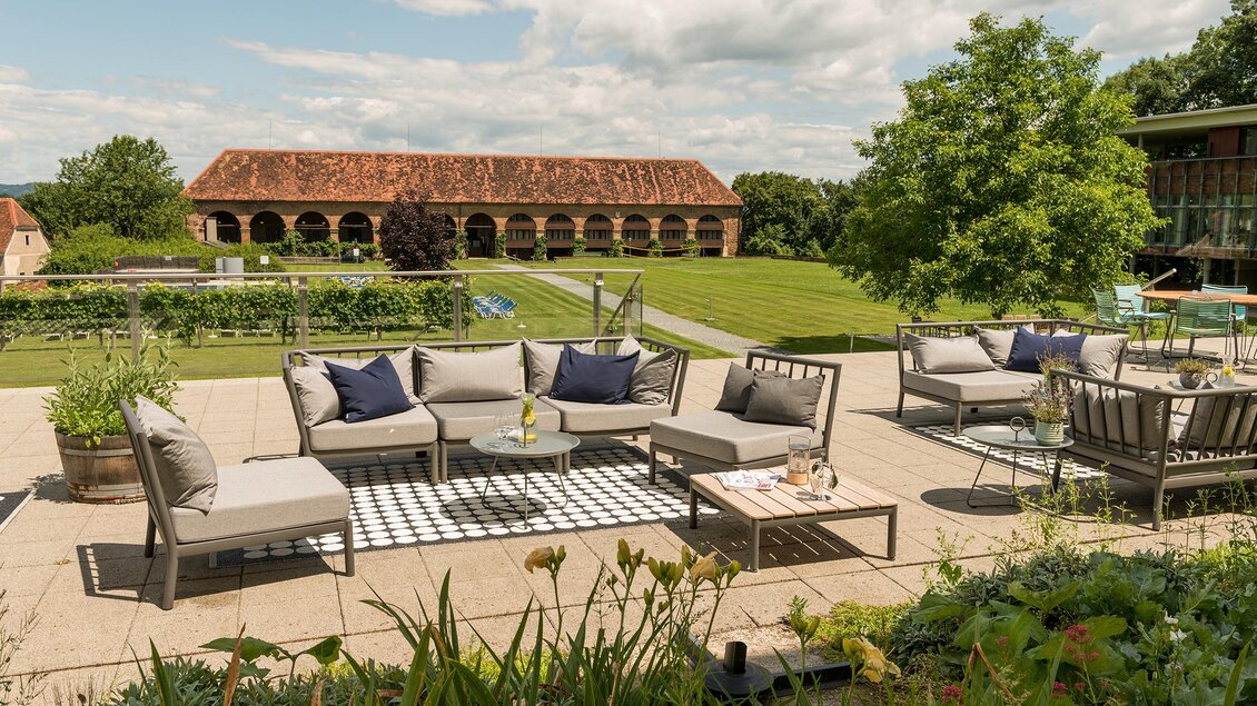 Eine einladende Terrasse mit modernen Gartenmöbeln und dekorativen Kissen. Im Hintergrund befindet sich ein großes, grün bewachsenes Grundstück mit einem historischen Gebäude. | © Stefan Kristoferitsch