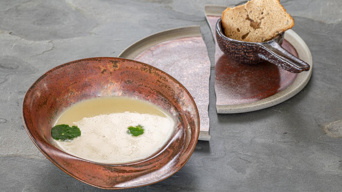 Eine aromatische Suppe in einer dekorativen Schüssel mit frischen Kräutern. Neben der Schüssel liegt ein kleiner Teller mit Brot. | © Schneebauer