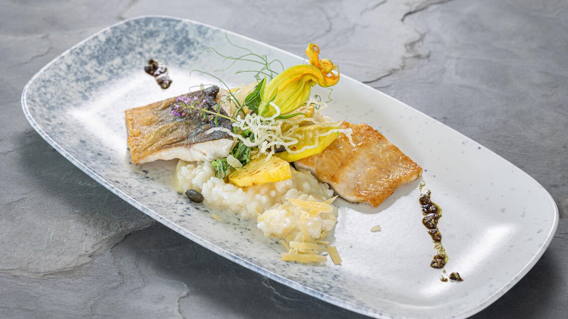 Ein eleganter Teller mit zwei perfekt zubereiteten Fischstücken, serviert auf einem Bett aus Risotto. Frisches Gemüse und essbare Blüten runden das Gericht visuell ab. | © Schneebauer