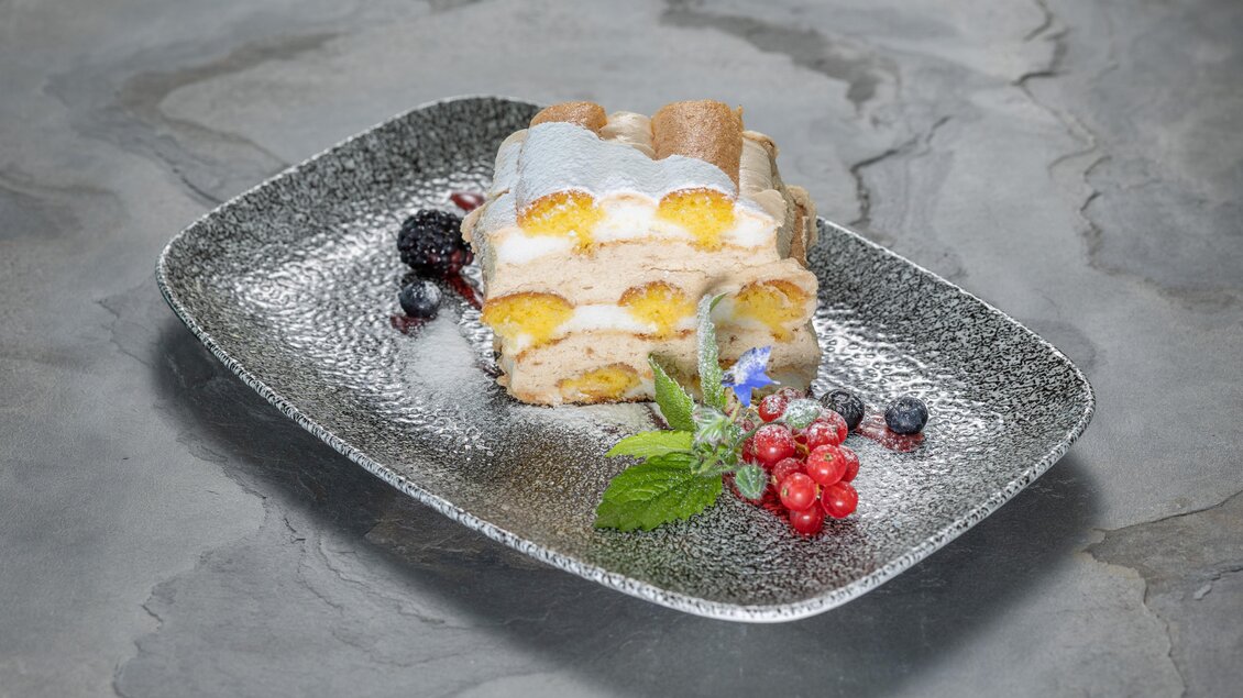 Ein köstliches Dessert auf einem schwarzen Teller, bestehend aus Löffelbiskuit und einer cremigen Füllung. Dekoriert mit frischen Beeren und einem Minzblatt. | © Schneebauer