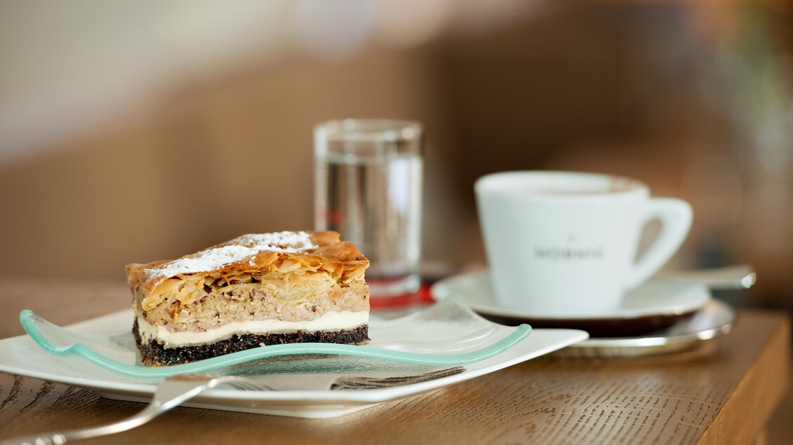 Ein Stück Kuchen auf einem Teller, daneben ein Glas Wasser und eine Tasse Kaffee. Der Kuchen hat eine schichtartige Struktur und ist mit Puderzucker bestreut. | © Stefan Kristoferitsch