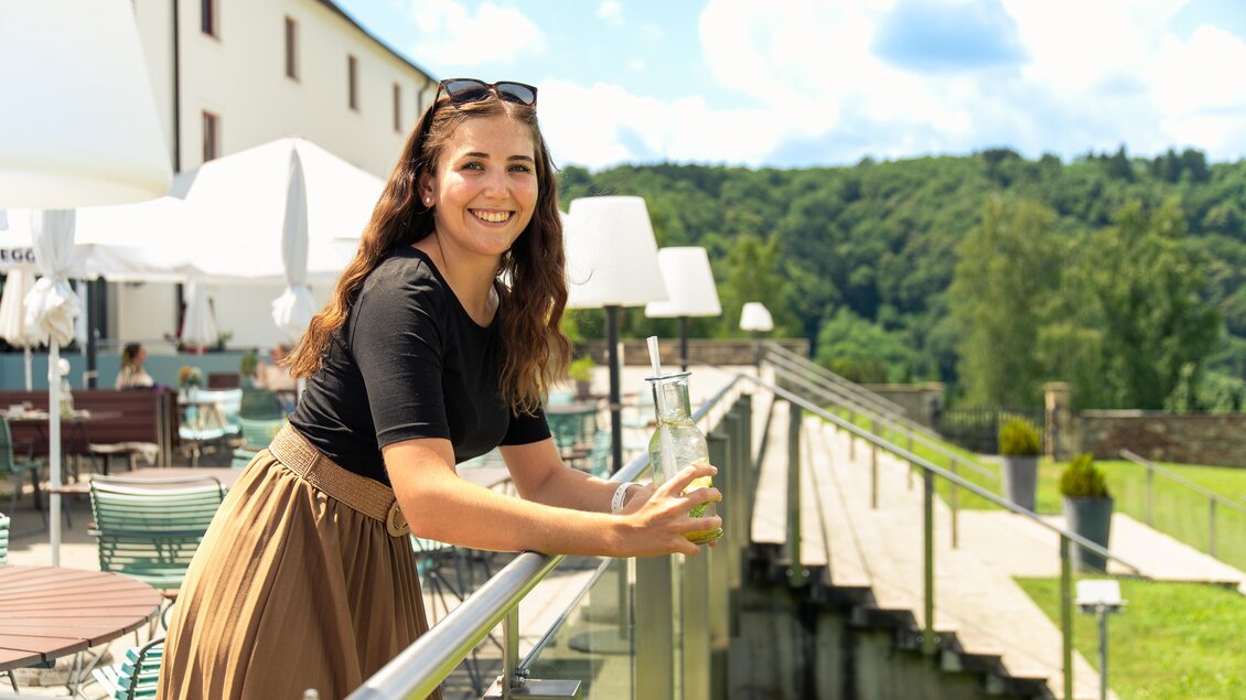 Eine junge Frau steht lächelnd an einem Geländer. Im Hintergrund sind Bäume und ein hellblauer Himmel zu sehen. | © Schlosstaverne Schloss Seggau
