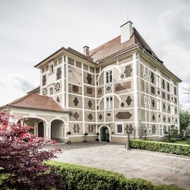 An elegant building with unique facade design. Surrounded by greenery and flowers, it appears inviting and charming. | © Schloss Farrach