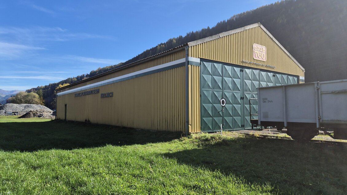 Ein großes gelbes Gebäude mit grünen Türen steht auf einer Wiese. Im Hintergrund sind Berge und blauer Himmel zu sehen. | © TVB Murau