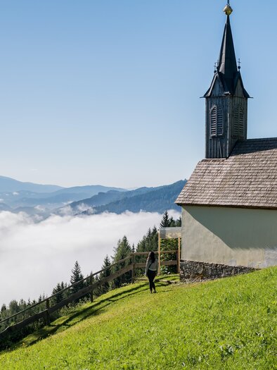 Eine kleine Kirche auf einer grünen Wiese mit Bäumen im Hintergrund. Über der Landschaft liegt ein Nebelteppich, während die Berge in der Ferne sichtbar sind. | © Anita Fössl