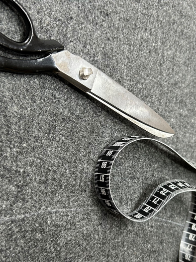 A pair of scissors lies on a gray background next to a tape measure. The tape measure is partially rolled up and shows numbers. | © Maria Greindl