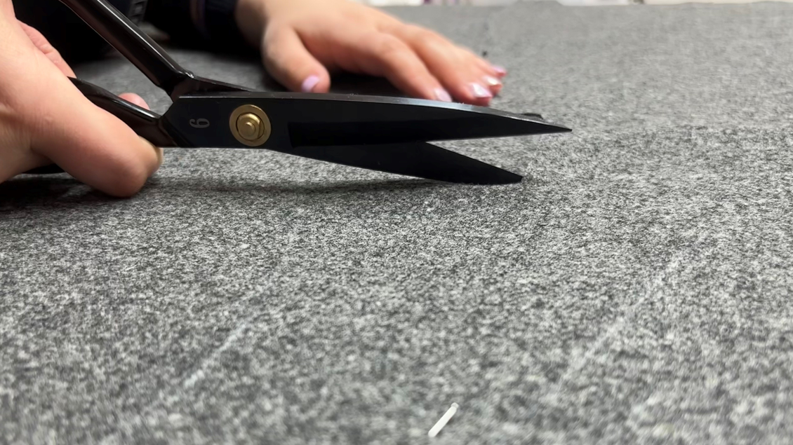 Eine Hand hält eine Schere über grauem Stoff. Der Fokus liegt auf den Schneidklingen der Schere. | © Maria Greindl