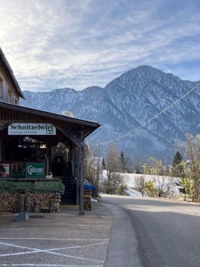 A cozy building with a bistro sign stands on a street. In the background, snowy mountains rise under a clear sky. | © TVB_Ausseerland Salzkammergut_Heike Temmel