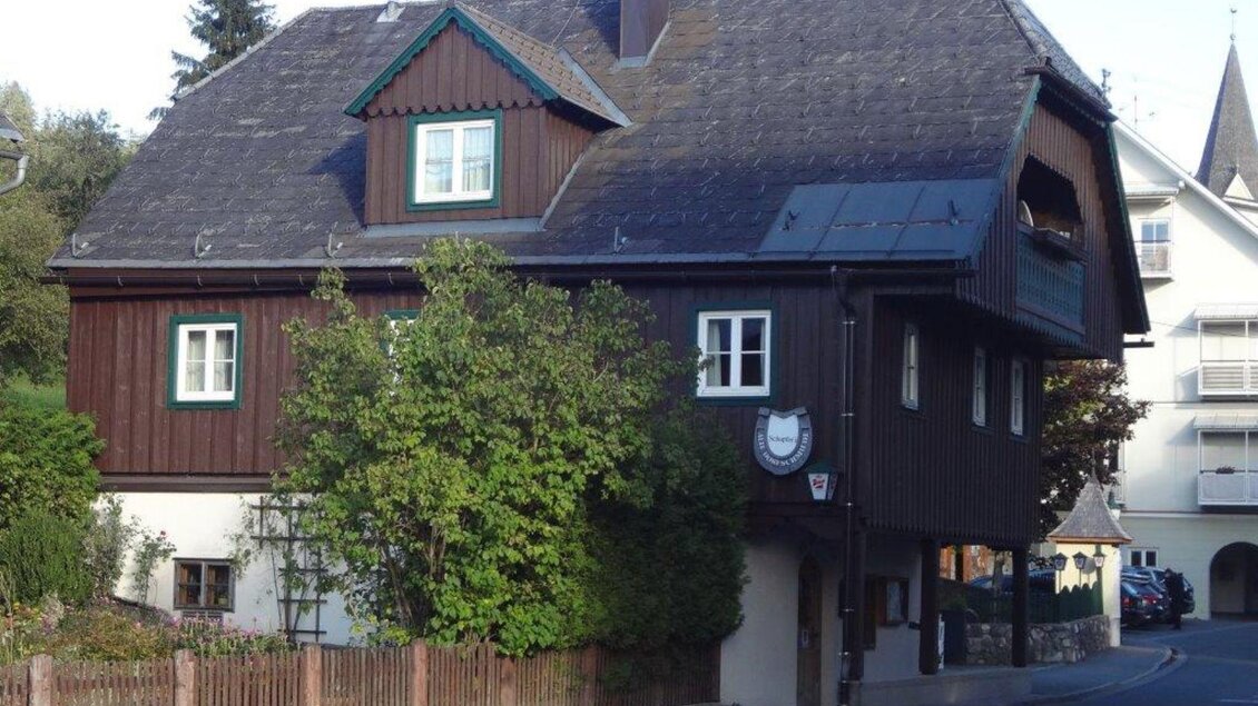 Ein traditionelles, dunkelbraunes Holzhaus mit mehreren Fenstern. Umgeben von Bäumen und einer kleinen Hecke in einer ruhigen Straße. | © Roland Schupfer