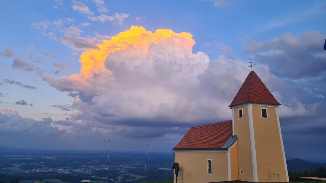 Eine kleine gelbe Kirche steht auf einem Hügel. Im Hintergrund sind beeindruckende Wolken und ein schöner Himmel zu sehen. | © Mesnerhaus