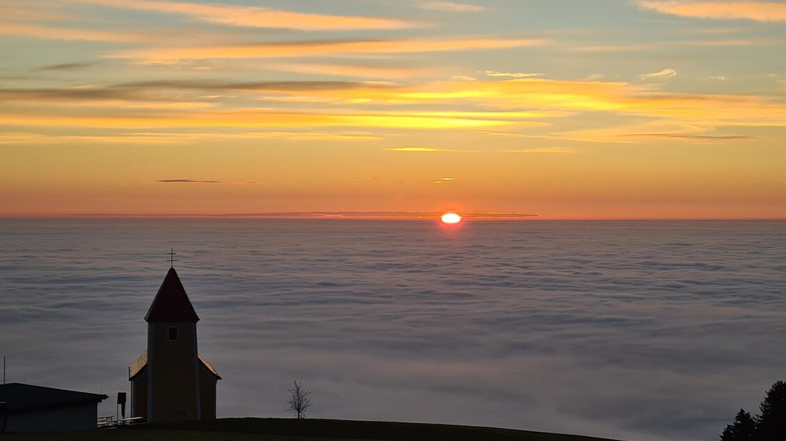 Eine kleine Kirche steht auf einem Hügel über dem Nebel. Der Sonnenuntergang färbt den Himmel in warmen Farben. | © Mesnerhaus
