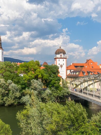 Eine malerische Stadtansicht mit einer Brücke über einen Fluss. Im Hintergrund sind historische Gebäude und eine Kirche zu sehen, umgeben von Bäumen und einem blauen Himmel. | © TVB Erzberg-Leoben