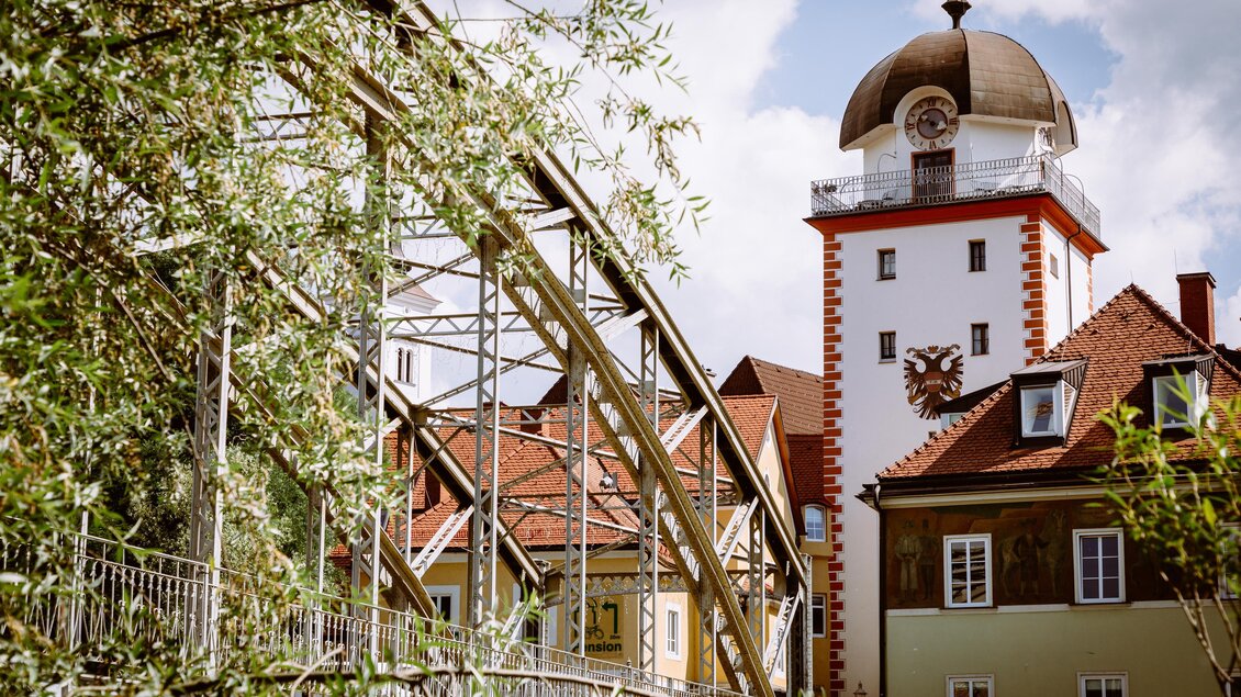 Eine malerische Stadtansicht mit einer alten Brücke und einem historischen Turm. Die Gebäude zeigen traditionelle Architektur unter einem blauen Himmel. | © TVB Erzberg-Leoben