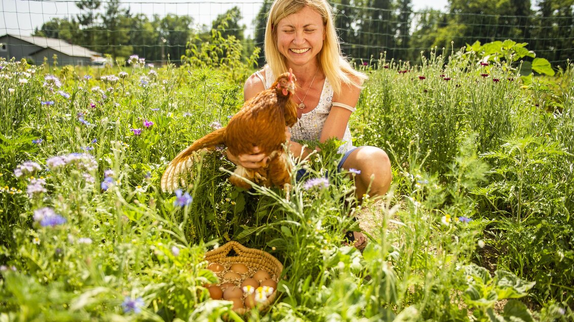 Eine Frau sitzt in einem blühenden Garten und hält ein Huhn in der Hand. Um sie herum sind bunte Blumen und eine natürliche Landschaft zu sehen. | © Ama Genussregion