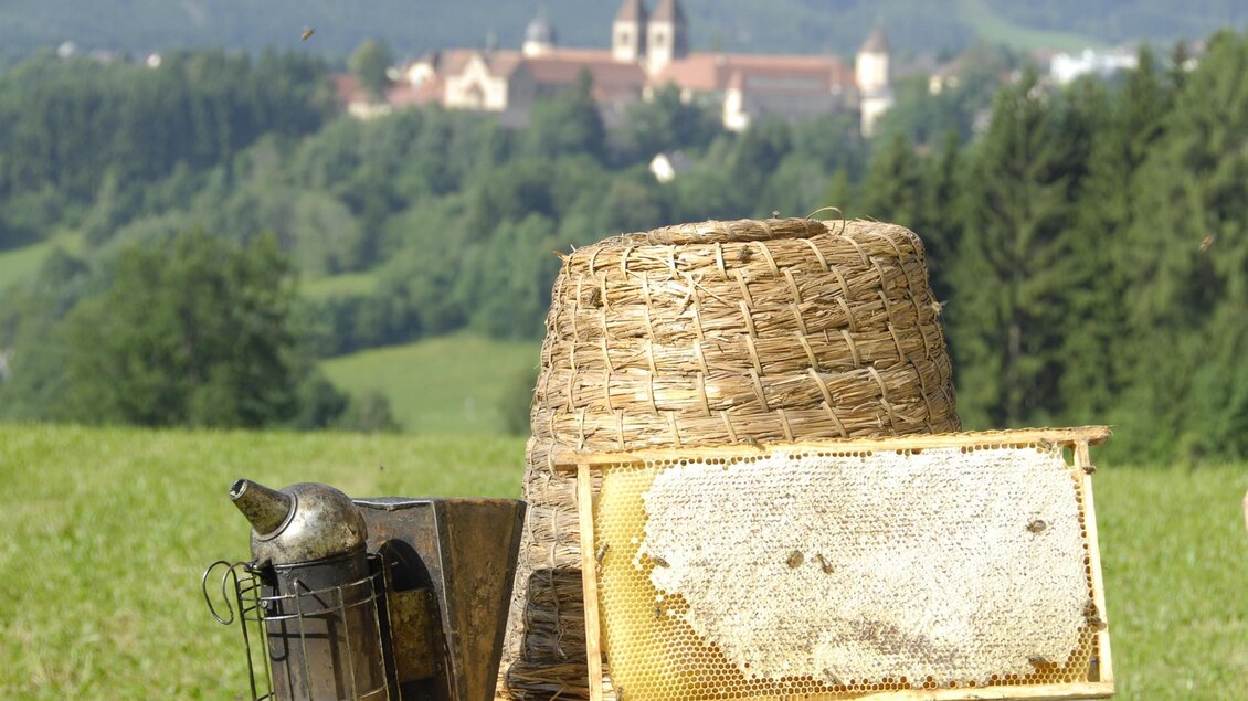Eine Imkerausrüstung steht auf einem Tisch, darunter ein Wabenrahmen mit Honig. Im Hintergrund sind grüne Hügel und ein historisches Gebäude zu sehen. | © Seckauer Waldhonig – Imkerei Wachter