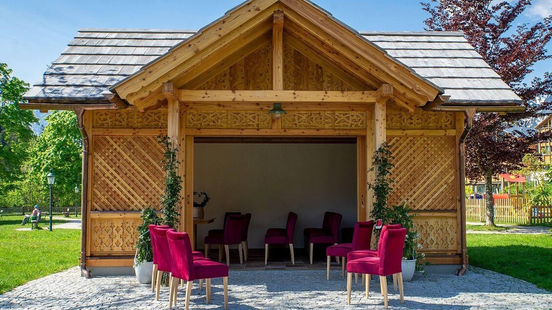 <p>Ein gemütliches Holzhaus mit einem offenen Eingangsbereich. Vor dem Haus stehen rote Stühle auf einem Pflasterboden.</p> | © Lebensbilder_Christine-Struz