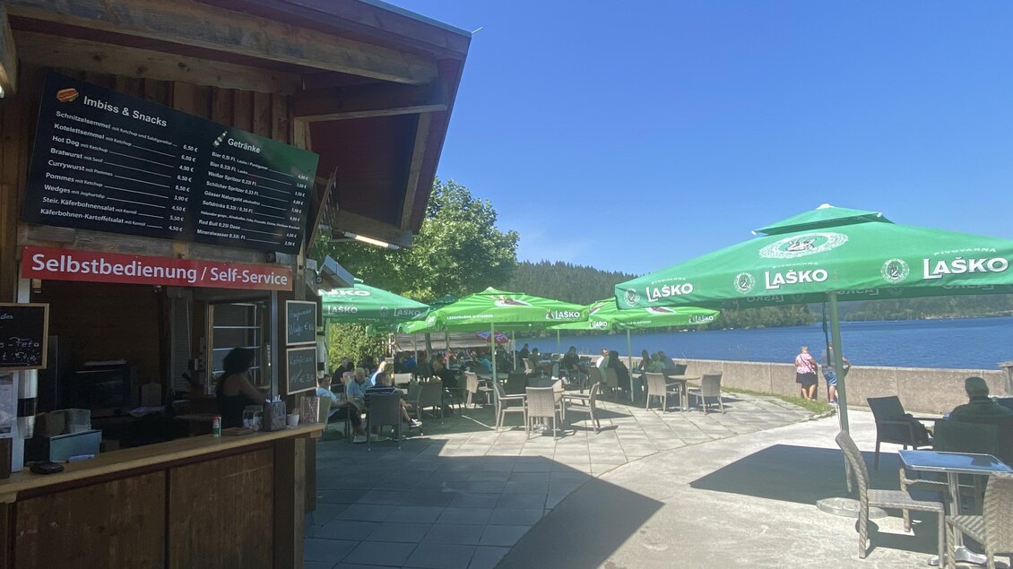 Ein gemütlicher Imbiss am Wasser mit Sonnenschirmen. Viele Besucher genießen die Aussicht und die entspannte Atmosphäre.
