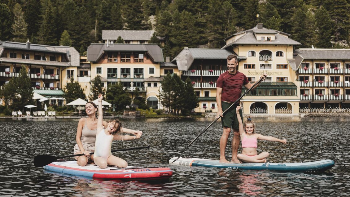 Eine Familie paddelt fröhlich auf Stand-Up-Paddle-Boards auf einem ruhigen See. Im Hintergrund sind malerische Gebäude und Bäume zu sehen. | © Seehotel Jägerwirt
