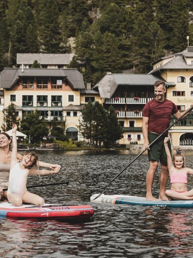 Eine Familie paddelt fröhlich auf Stand-Up-Paddle-Boards auf einem ruhigen See. Im Hintergrund sind malerische Gebäude und Bäume zu sehen. | © Seehotel Jägerwirt