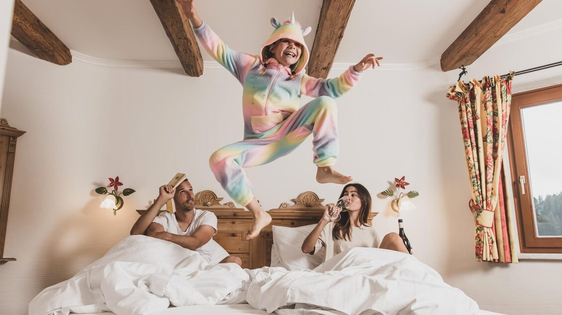 Drei Personen in einem Schlafzimmer haben Spaß. Eine Person springt im Einhorn-Schlafanzug auf dem Bett, während die anderen entspannt liegen und Snacks genießen. | © Seehotel Jägerwirt