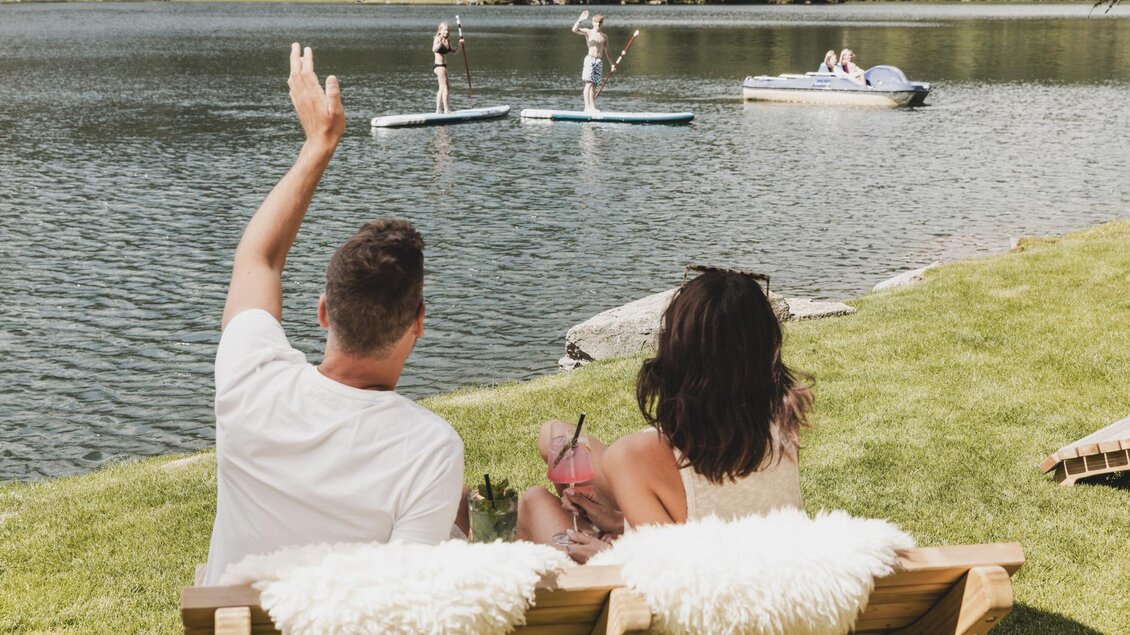 Ein Paar sitzt auf einer Bank am Wasser und genießt die Aussicht. Im Hintergrund stehen Personen auf Stand-Up-Paddleboards. | © Seehotel Jägerwirt