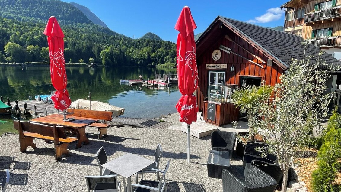 Ein gemütlicher Außenbereich am See mit Tischen, Stühlen und Sonnenschirmen. Im Hintergrund sind grüne Hügel und ein Klärkanal zu sehen. | © TVB Ausseerland/Bettina Scheck