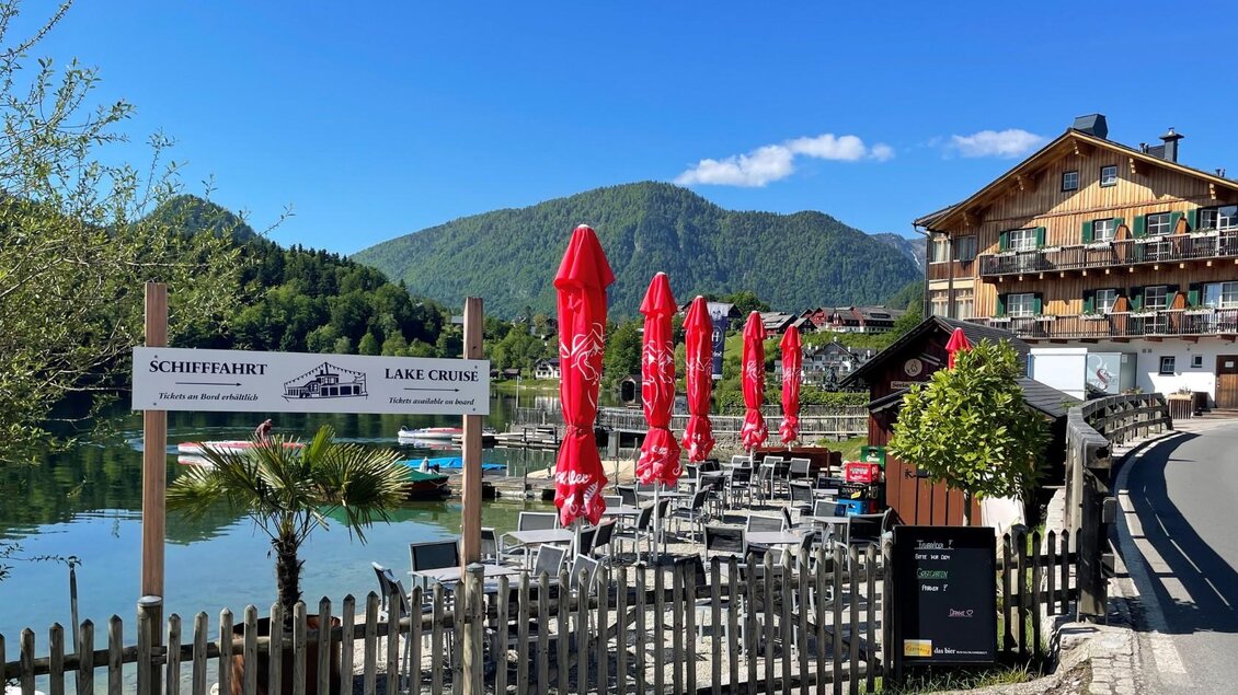 Ein schöner See mit roten Sonnenschirmen und gemütlichen Sitzplätzen. Im Hintergrund sind grüne Berge und ein charmantes Gebäude zu sehen. | © TVB Ausseerland/Bettina Scheck