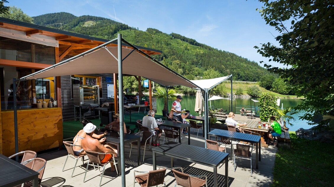 Ein einladendes Café am Wasser mit Terrassen und mehreren Gästen. Im Hintergrund sind grüne Hügel und ein klarer Himmel zu sehen. | © TV Region Graz - René Vidalli