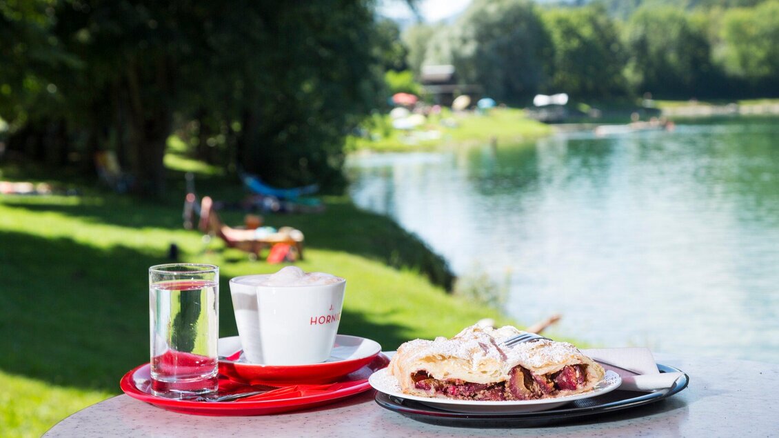 Ein Tisch am Wasser mit einer Tasse Kaffee, einem Glas Wasser und einem Stück Kuchen. Im Hintergrund sind grüne Wiesen und eine ruhige Wasseroberfläche zu sehen. | © TV Region Graz - René Vidalli