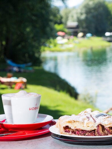 A table by the water with a cup of coffee, a glass of water, and a piece of cake. In the background, green meadows and a calm surface of the water can be seen. | © TV Region Graz - René Vidalli