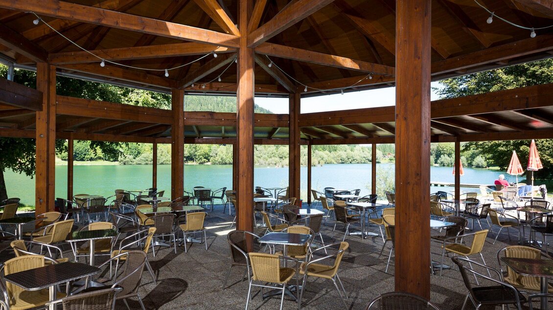Ein gemütlicher Restaurantbereich mit Holzdecken und Tischen am Wasser. Der Blick fällt auf einen ruhigen See und umgebende Natur. | © TV Region Graz- René Vidalli