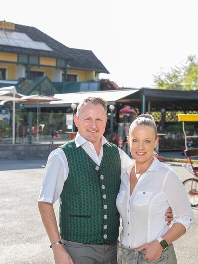 A couple is standing smiling in front of a restaurant with a sunny outdoor terrace. In the background, pedal boats and bicycles can be seen. | © Seerestaurant Gruber
