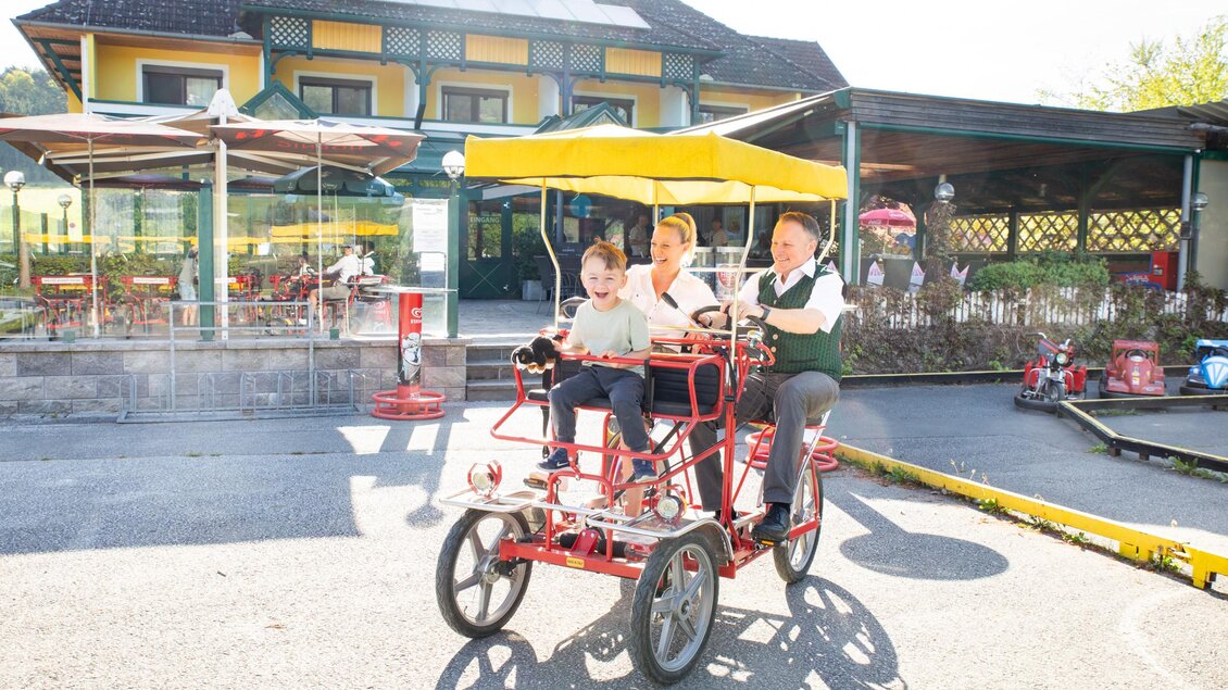 Eine Familie fährt mit einem lustigen Dreirad vor einem Restaurant. Im Hintergrund sind Sitzbereiche im Freien und sonniges Wetter zu sehen. | © Seerestaurant Gruber
