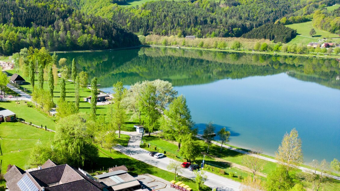 Ein ruhiger See umgeben von grünen Hügeln und Bäumen. Im Vordergrund sind Gebäude und Wege sichtbar. | © Seerestaurant Gruber