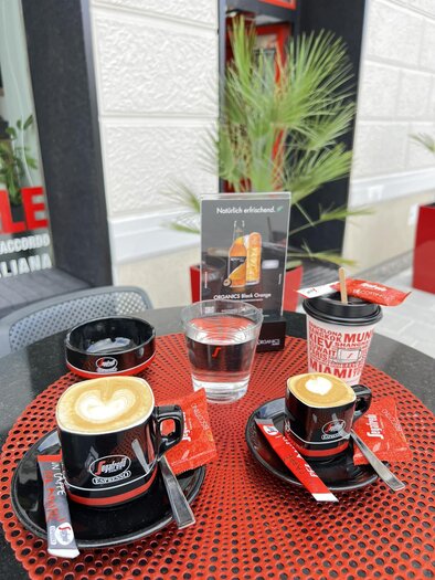 Ein Tisch mit zwei Tassen Kaffee, einem Wasserglas und einem Getränkebecher. Im Hintergrund ist eine Pflanze und ein Schild sichtbar. | © Andi Bäuchl