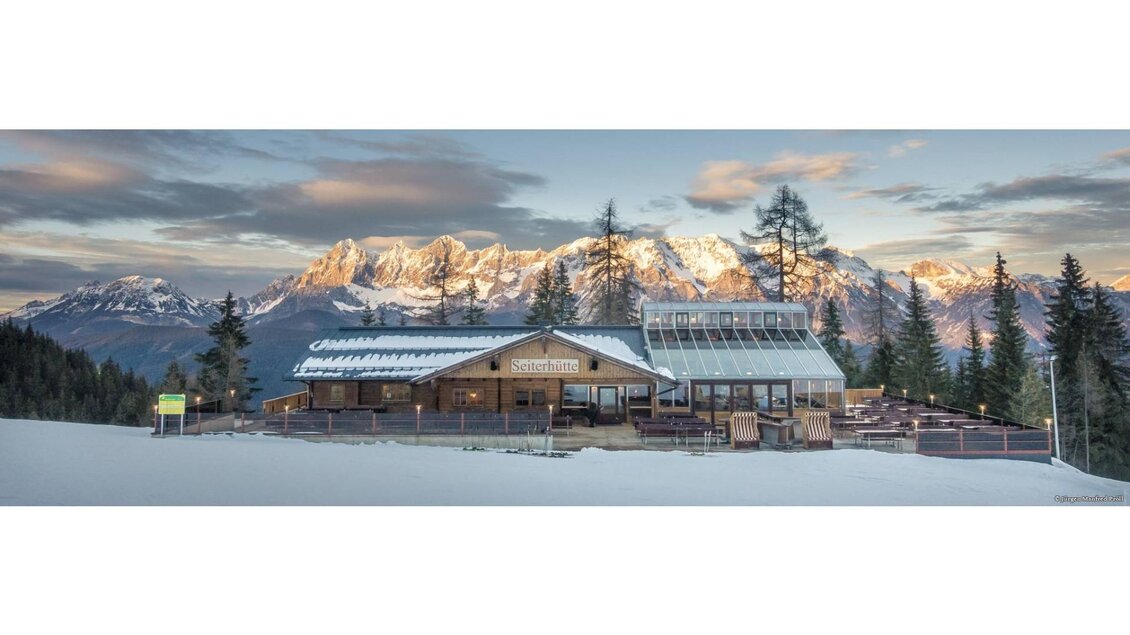 Ein charmantes Berghaus mit einer großen Terrasse, umgeben von schneebedeckten Bergen und Nadelbäumen. Der Himmel ist klar und die Bergspitzen sind in warmes Licht getaucht. | © Jürgen Manfred Pröll
