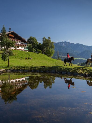 A picturesque landscape with a mountain house, surrounded by green meadows. Two riders are reflected in the calm surface of a pond.