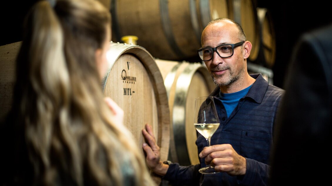 Ein Mann mit Brille hält ein Glas Wein und spricht mit einer Frau. Im Hintergrund sind Holzfässer zu sehen, die typisch für eine Weinkellerei sind. | © Stephan Friesinger