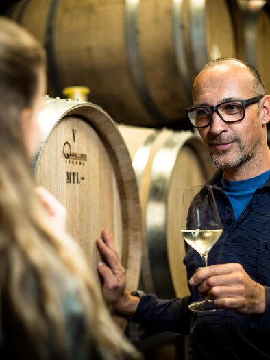 A man with glasses is holding a glass of wine and talking to a woman. In the background, wooden barrels typical of a wine cellar can be seen. | © Stephan Friesinger