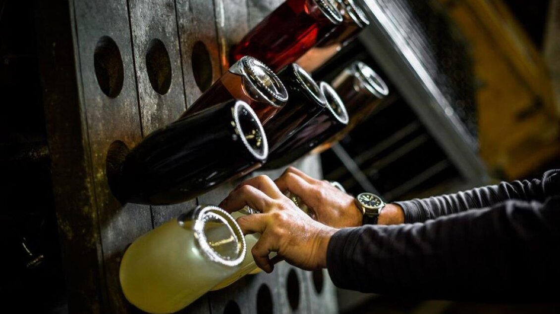 Eine Hand hält eine Flasche, während mehrere andere Flaschen auf einem Rack stehen. Die Flaschen sind in verschiedenen Farben und stehen senkrecht. | © Stephan Friesinger